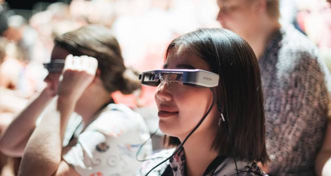 photo avec au premier plan une jeune femme portant des lunettes reliées à un fil, en train de regarder un spectacle. Au second plan, deux autres personnes dont une a aussi ces lunettes