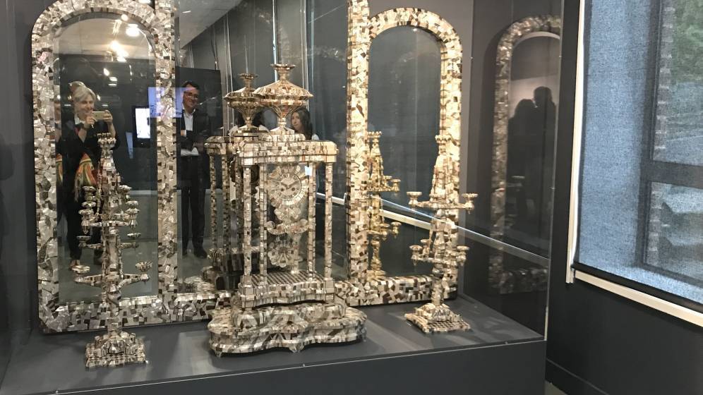 Vue d’ensemble d’une vitrine d’exposition du musée de la Nacre et de la Tabletterie à Méru. Au centre, un monumental autel en nacre finement sculptée, composé d’éléments architecturaux et ornementaux, avec colonnes, baldaquin et candélabres. L’ensemble est présenté dans une vitrine vitrée avec des miroirs en fond, amplifiant l’effet visuel. Une femme prenant une photo est visible, reflétée dans le miroir central. La scène est éclairée par des spots au plafond et baignée d’une lumière tamisée.