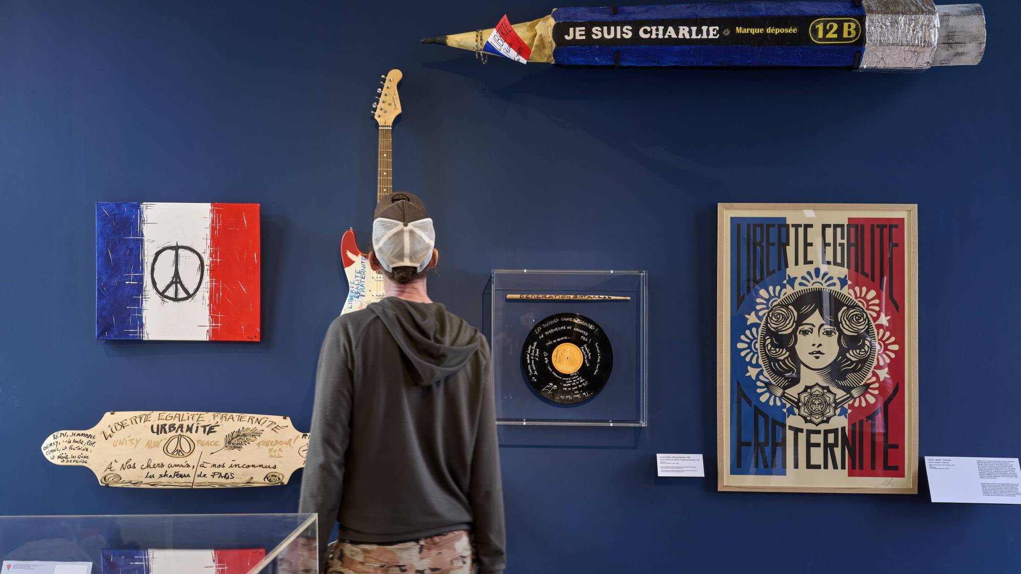 photo d'une personne de dos en train de regarder des objets accrochés à un mur : des affiches, un grand crayon, un vinyle