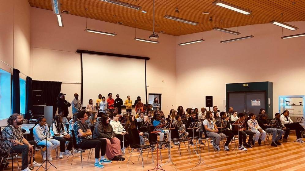 Dans une salle de répétition, les élèves de 2 classes orchestre sont assis et attendent de se mettre à jouer. Les élèves, un brin intimidés, font connaissance.