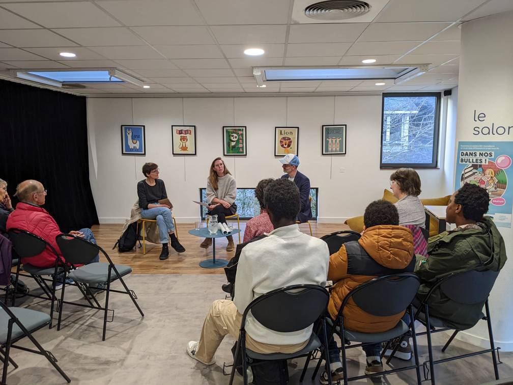 photo avec une petite conférence dans une bibliothèque avec du public de dos et trois intervenants de face assis
