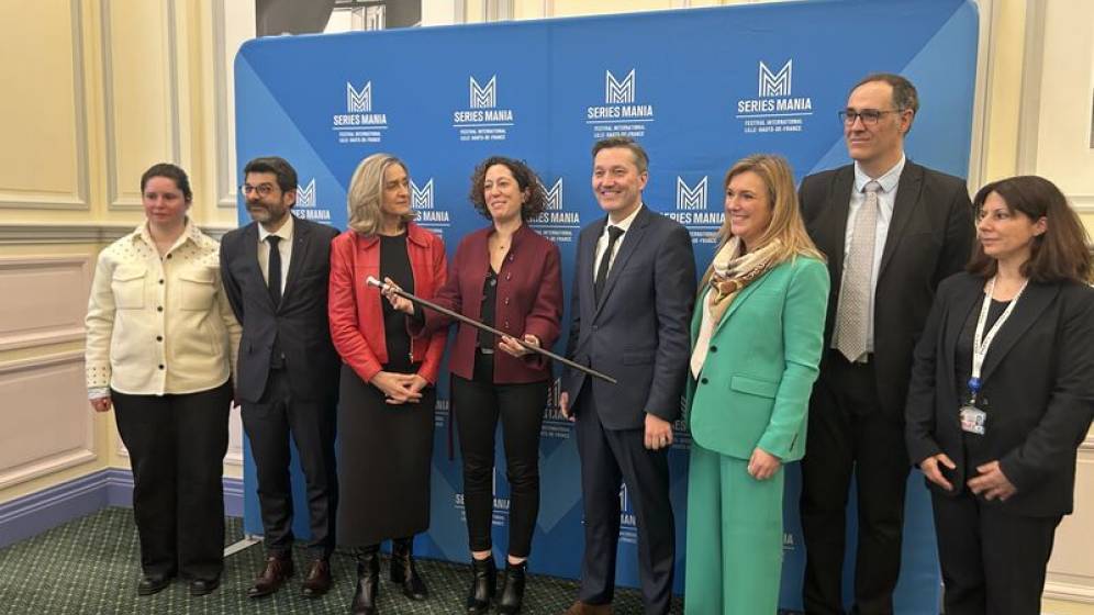 Un groupe de huit personnes pose devant un fond bleu avec le logo 'Séries Mania'. Les individus sont habillés de manière professionnelle, certains en costume, d'autres en tenues décontractées mais soignées. Ils participent à une séance photo avec le trophée dans la main de la femme au milieu, avec des expressions souriantes et engageantes.