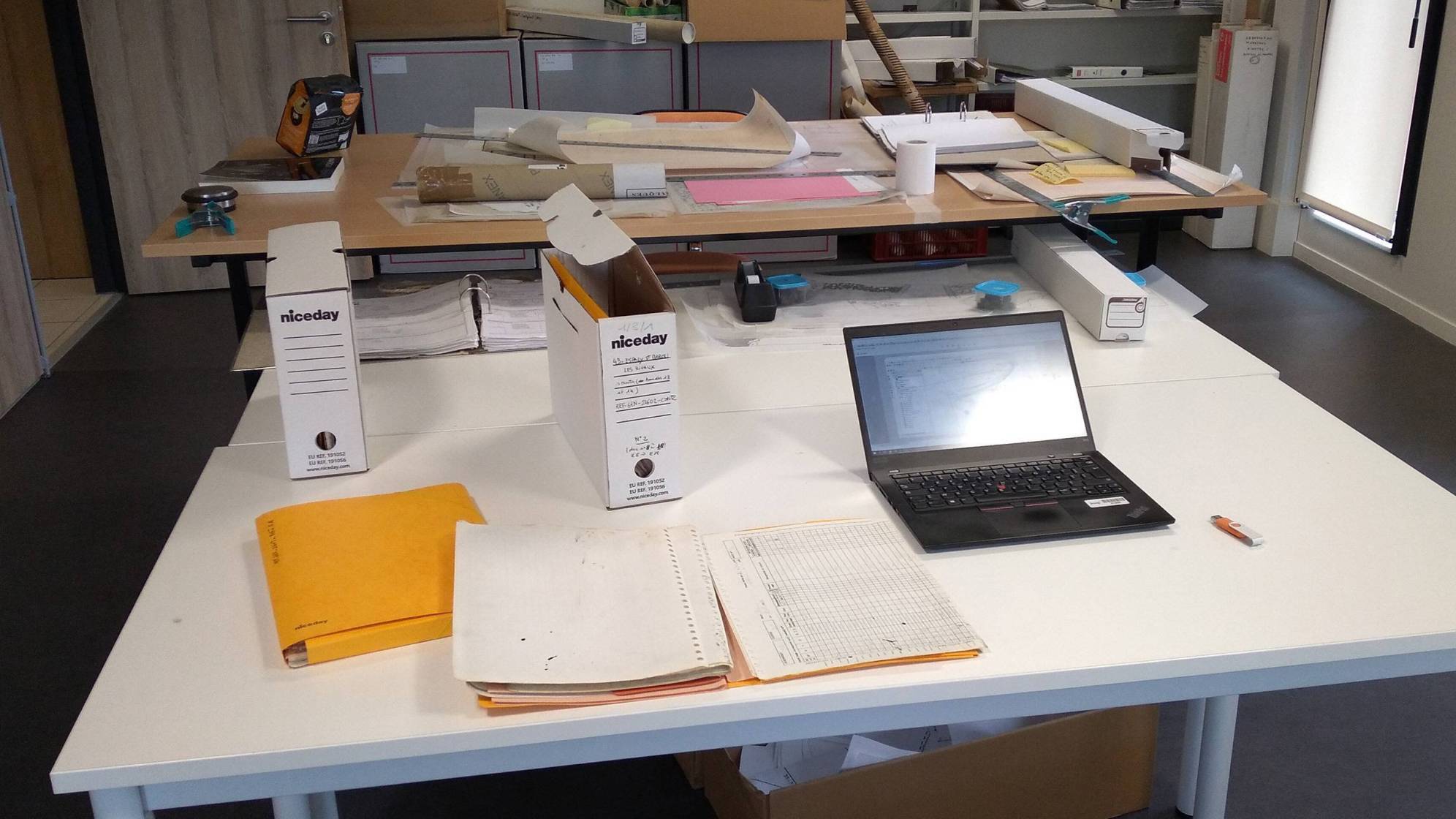 Sur des tables sont étalés des plans, des boîtes d'archives et des fiches d'enregistrement et un ordinateur ouvert. Sous la table, un grand cartons dans lesquels sont jetés tous les brouillons.