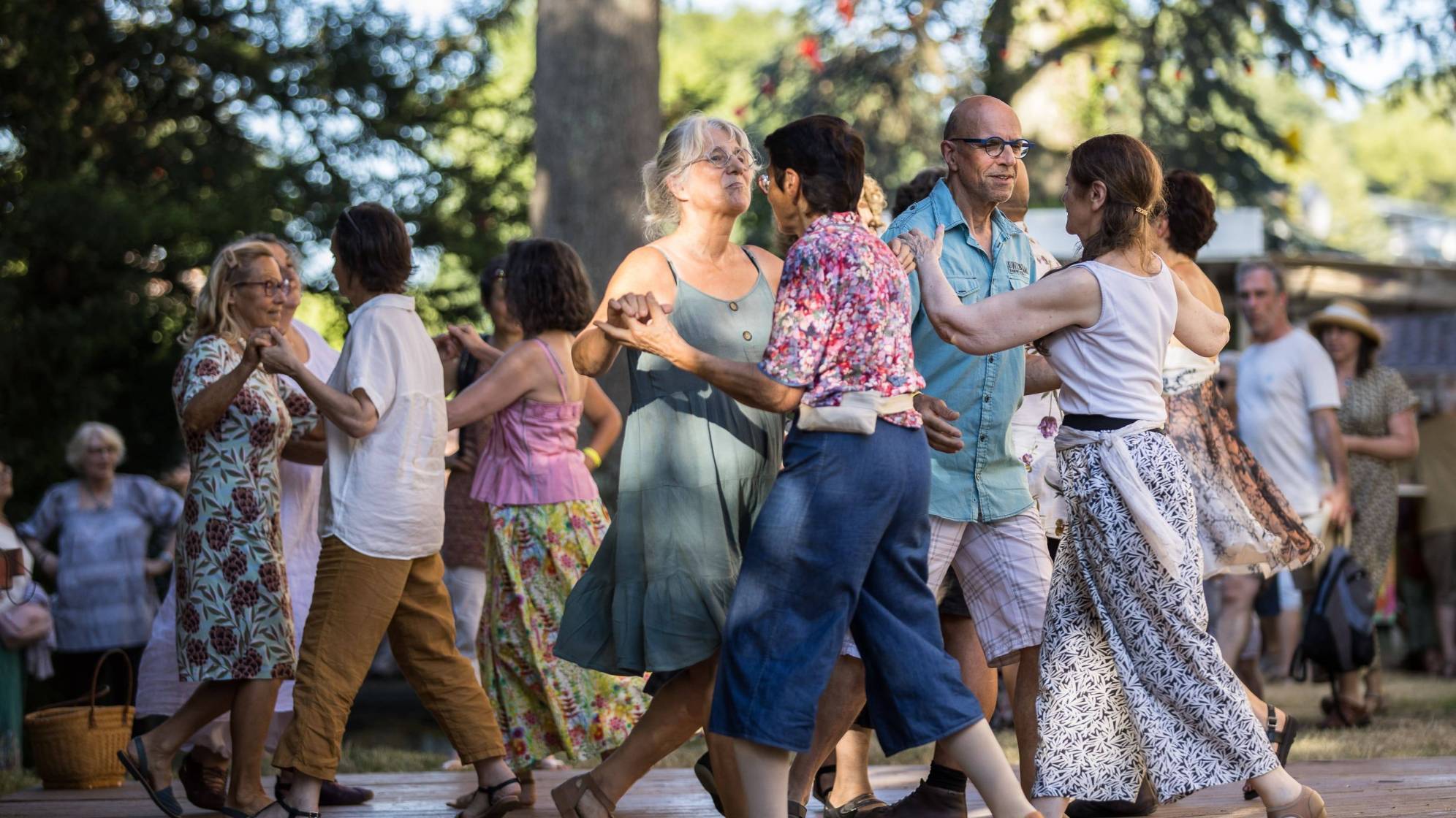 Des couples de genres et âges divers danses sur une scène de planche dans un décor de pins.
