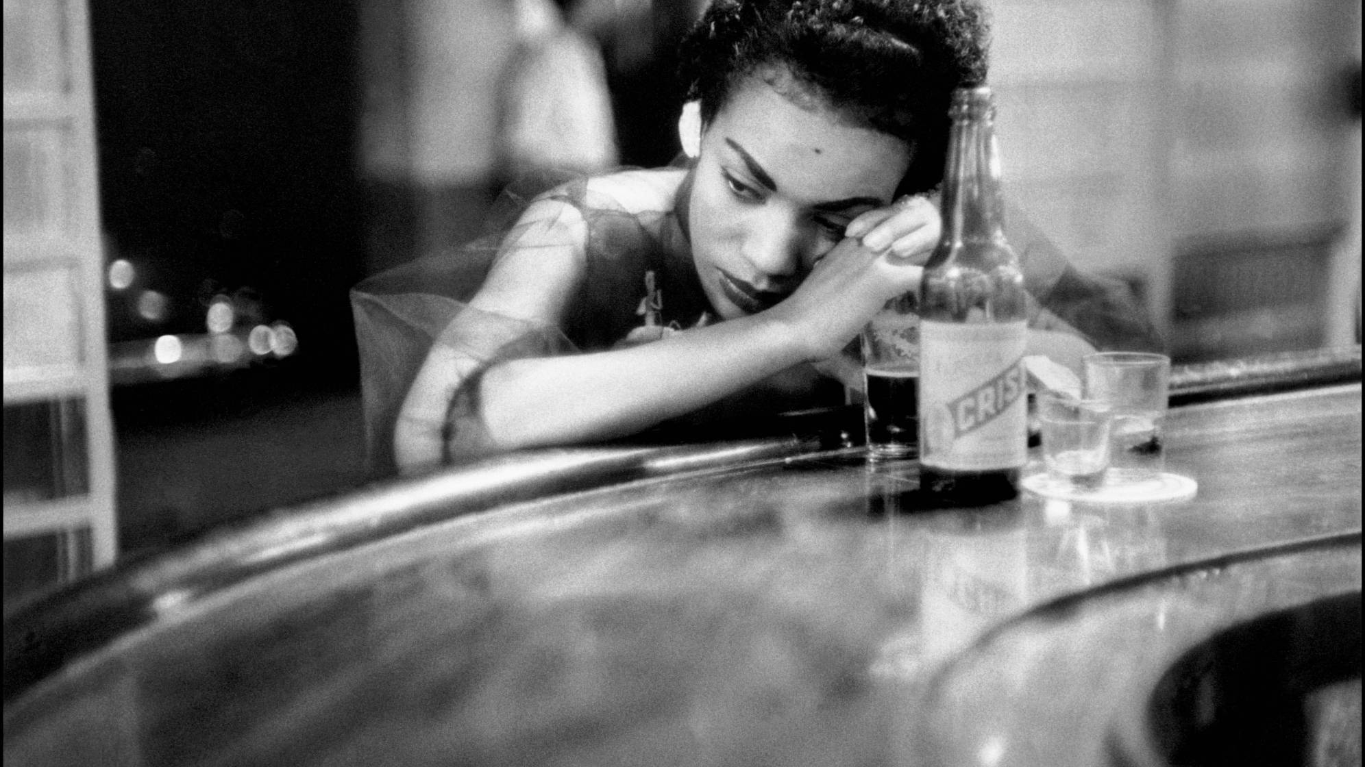 photo en noir et blanc d'Eve Arnold d'une femme accoudé à un bar avec une bouteille et un verre et qui regarde sur le côté, l'air songeur
