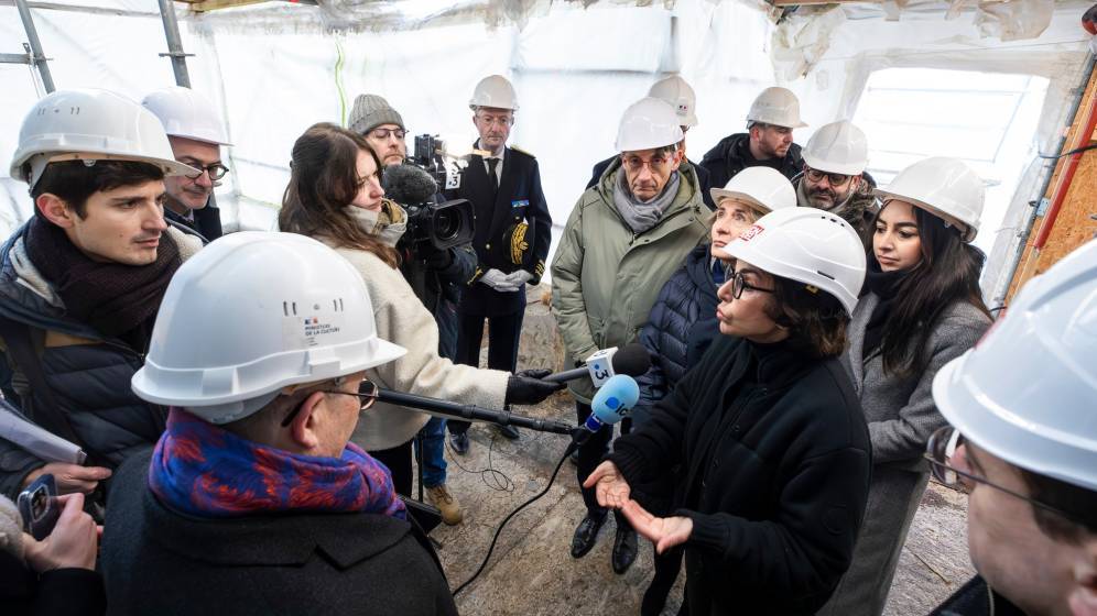 La ministre de la Culture, vêtue de noir et portant un casque de sécurité blanc, s'exprime face à un groupe de journalistes et d'officiels sur le chantier de restauration de la cathédrale Saint-Pierre de Beauvais. Des micros et des caméras sont dirigés vers elle, tandis que plusieurs personnes l'écoutent attentivement sous une structure temporaire protégeant le site en hauteur.