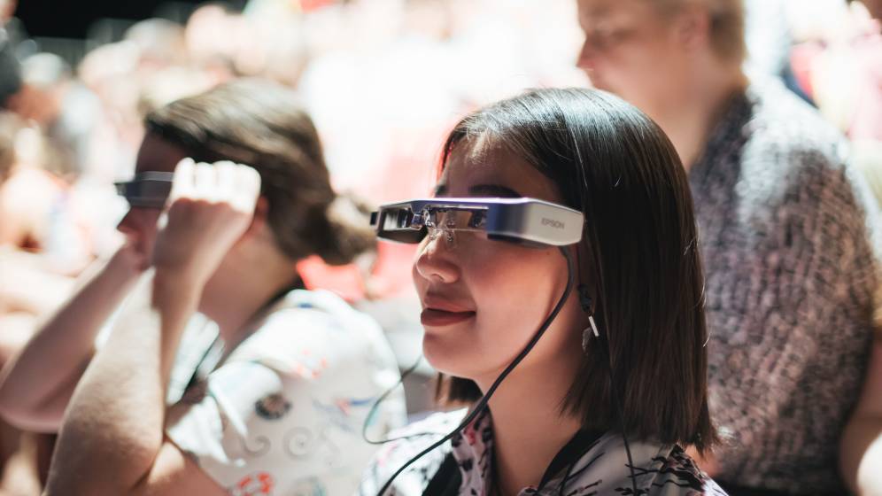 photo avec au premier plan une jeune femme portant des lunettes reliées à un fil, en train de regarder un spectacle. Au second plan, deux autres personnes dont une a aussi ces lunettes